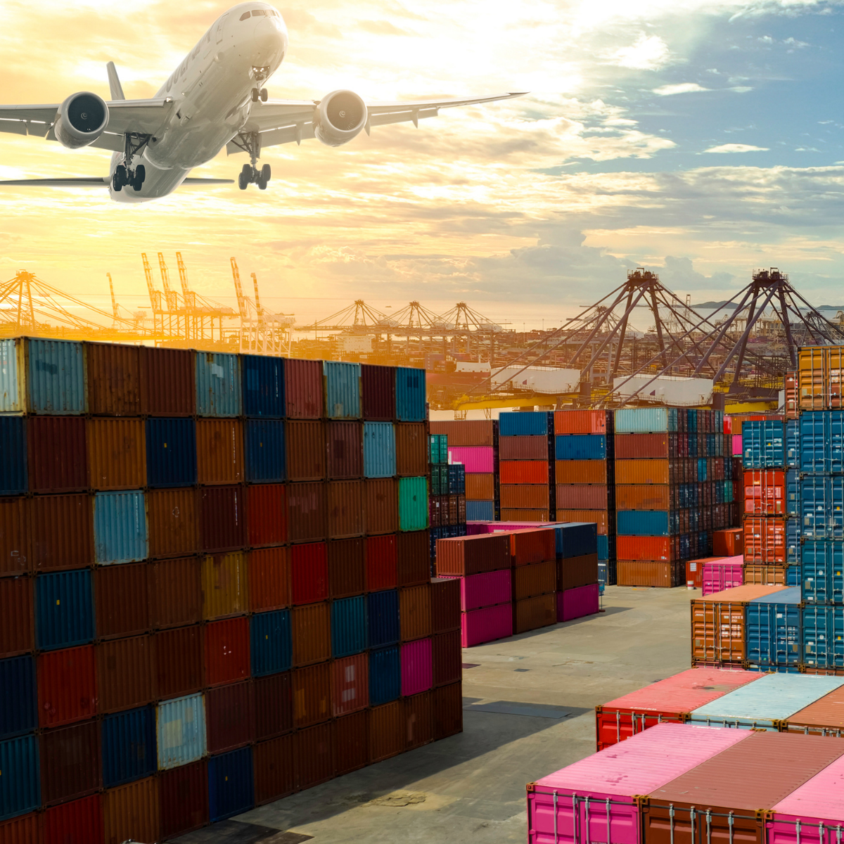 Een vrachtvliegtuig vliegt over een groot containerterrein met kleurrijke stapels zeecontainers bij zonsondergang.